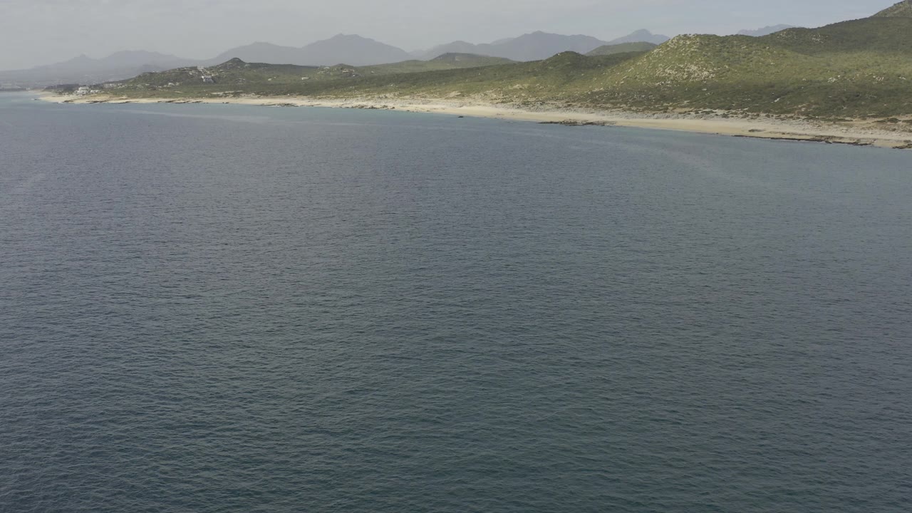 Aerial view in Baja California Sur, Mexico looking over dark blue water toward a long sandy beach, green coastal hills, hazy mountains, and no visible people.