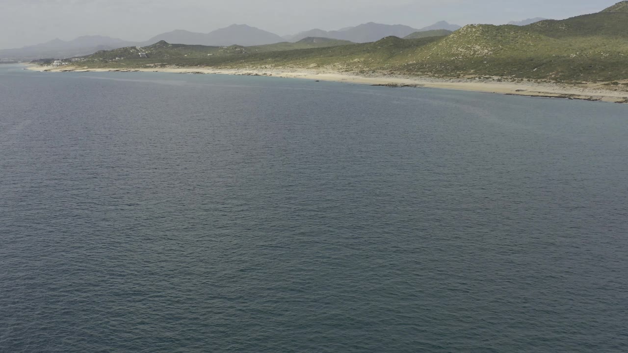 Wide aerial frame in Baja California Sur, Mexico with ocean covering most of the foreground, a pale beach across the middle, low green hills, distant mountains, and no visible people.