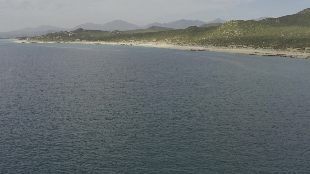 Baja California Sur coastline seen from above, with textured ocean water below, a narrow sandy beach, scrub-covered hills, layered mountains, and no visible people.