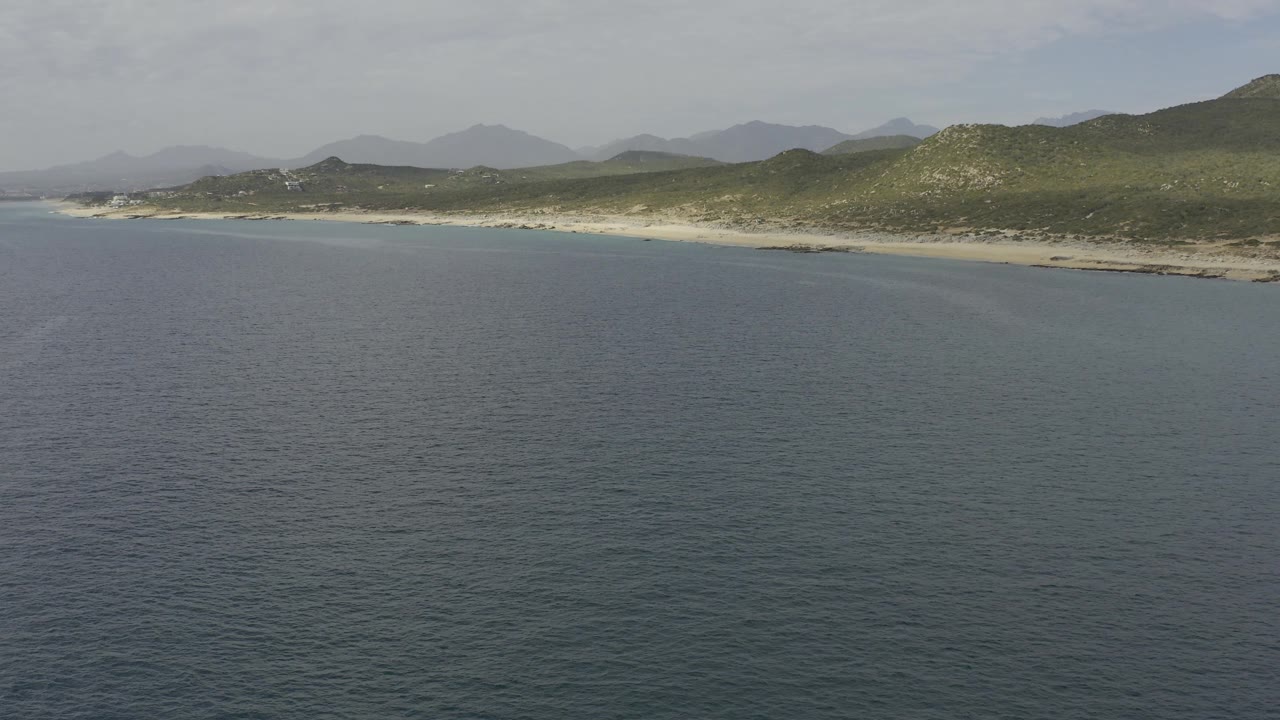 Aerial coastal view in Baja California Sur, Mexico showing muted blue water, a gently curving sandy shoreline, green hills with rocky patches, hazy mountains, and no visible people.