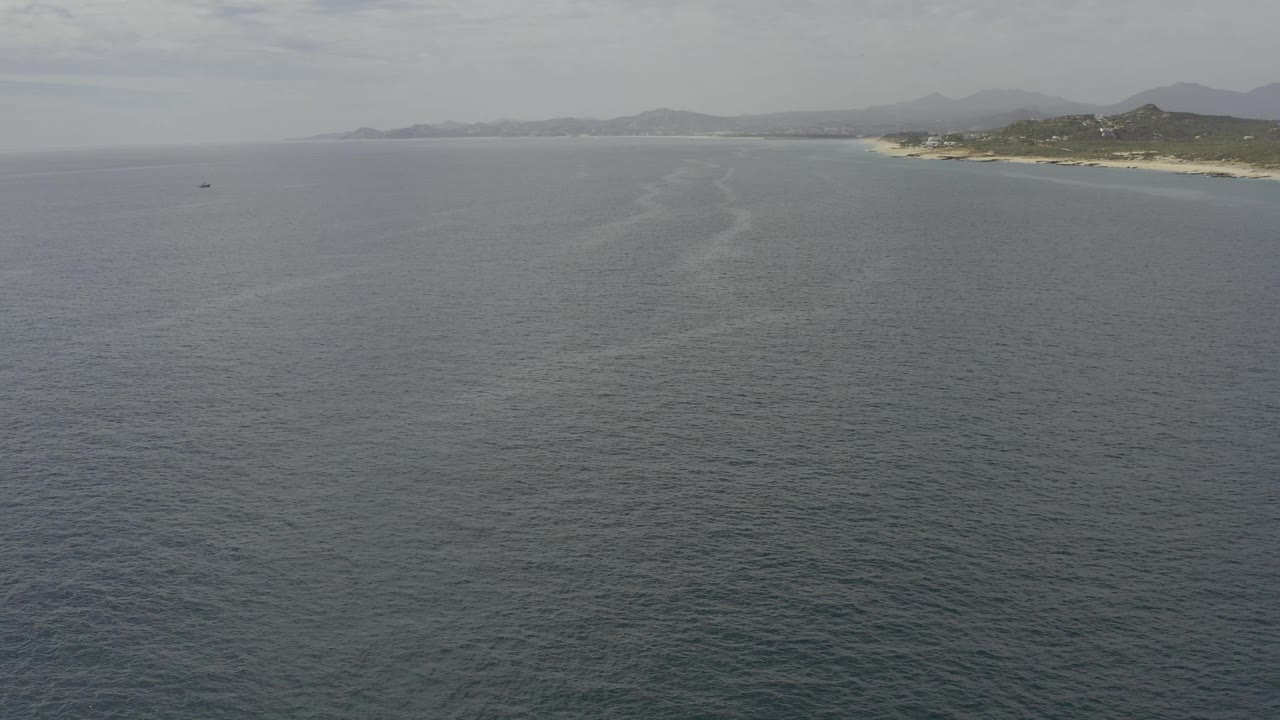 Wide aerial view in Baja California Sur with rippled ocean covering most of the frame, a light streak through the water, coastline at upper right, hazy hills, and no visible people.