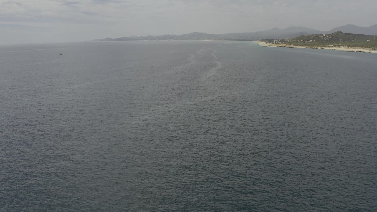 Aerial Baja California Sur coastal view with a curved pale wake or current line through the ocean, sandy shoreline to the right, distant headland to the left, and no visible people.
