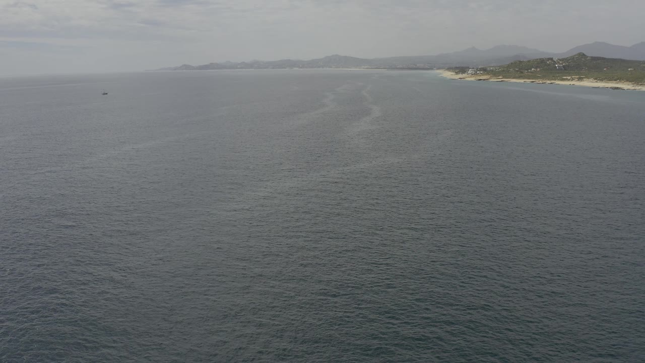 Aerial shot over Baja California Sur ocean with darker ripples in the foreground, a small dark speck on the water to the left, shoreline and hills near the horizon, and no visible people.