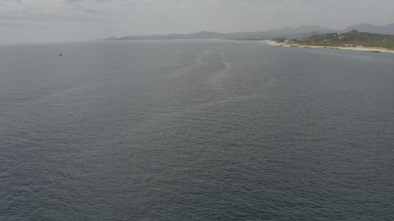 Opening aerial frame over gray-blue ocean off Baja California Sur, Mexico, with a sandy coastline and low hills along the upper right and no visible people.