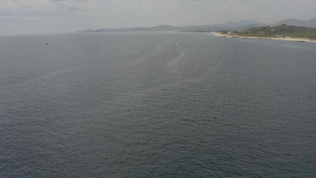 Mid-clip aerial frame over Baja California Sur water shows faint surface lines across the ocean, a narrow pale beach on the upper right, hazy hills beyond, and no visible people.
