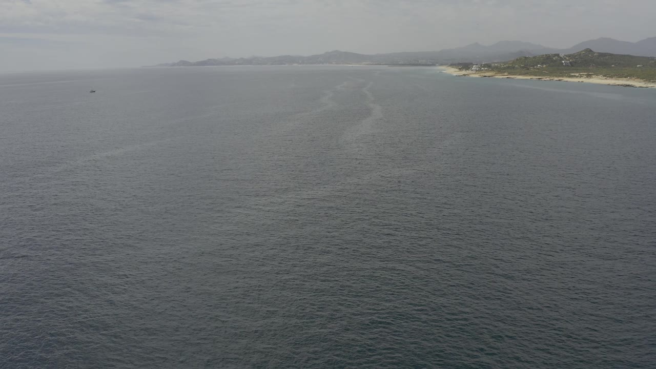Aerial frame from Baja California Sur with a broad field of muted ocean in the foreground, a hazy sky above, distant sandy shoreline to the right, and no visible people.