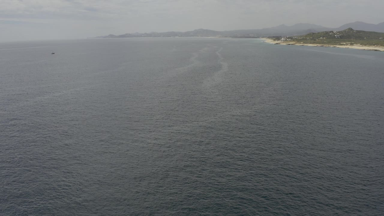 Wide aerial frame over gray-blue water along the Baja California Sur coastline, with pale beach and low hills in the distance and no visible people.