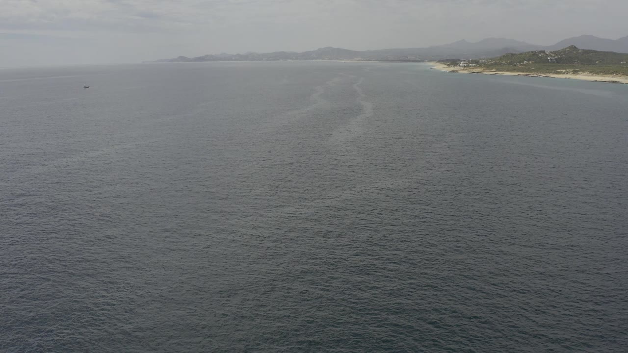 Aerial view of open water off Baja California Sur, with a pale shoreline curving at right, hazy mountains beyond, and no visible people.