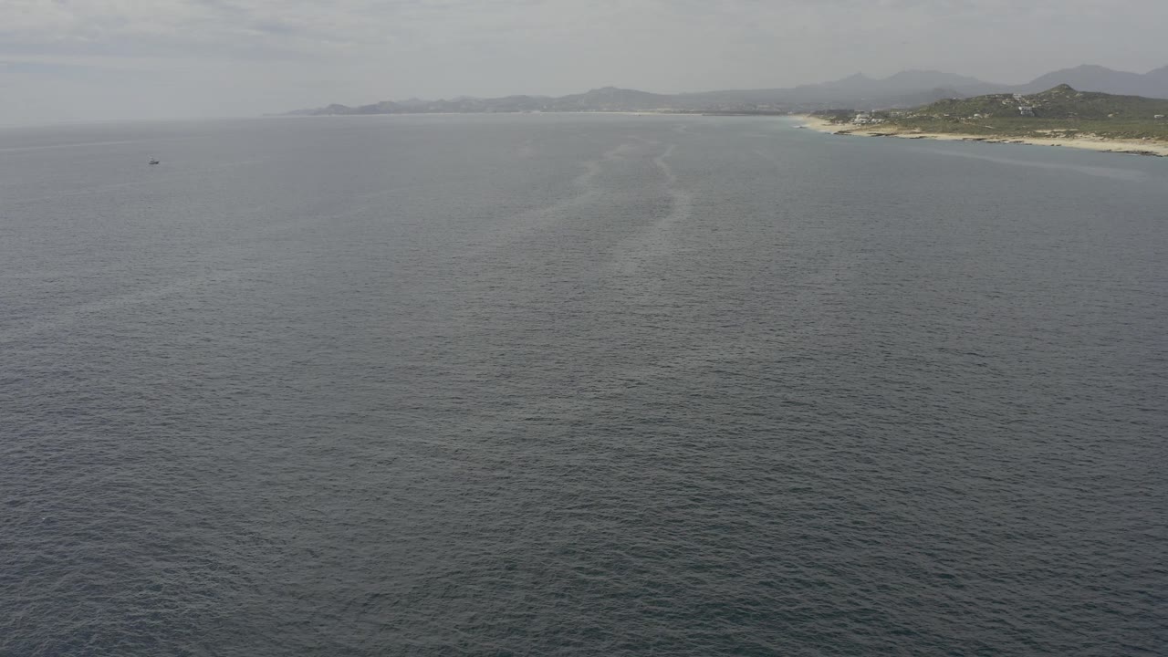Wide aerial frame over textured gray-blue ocean in Baja California Sur, with a faint foam or current line pointing toward the distant sandy coast and no visible people.