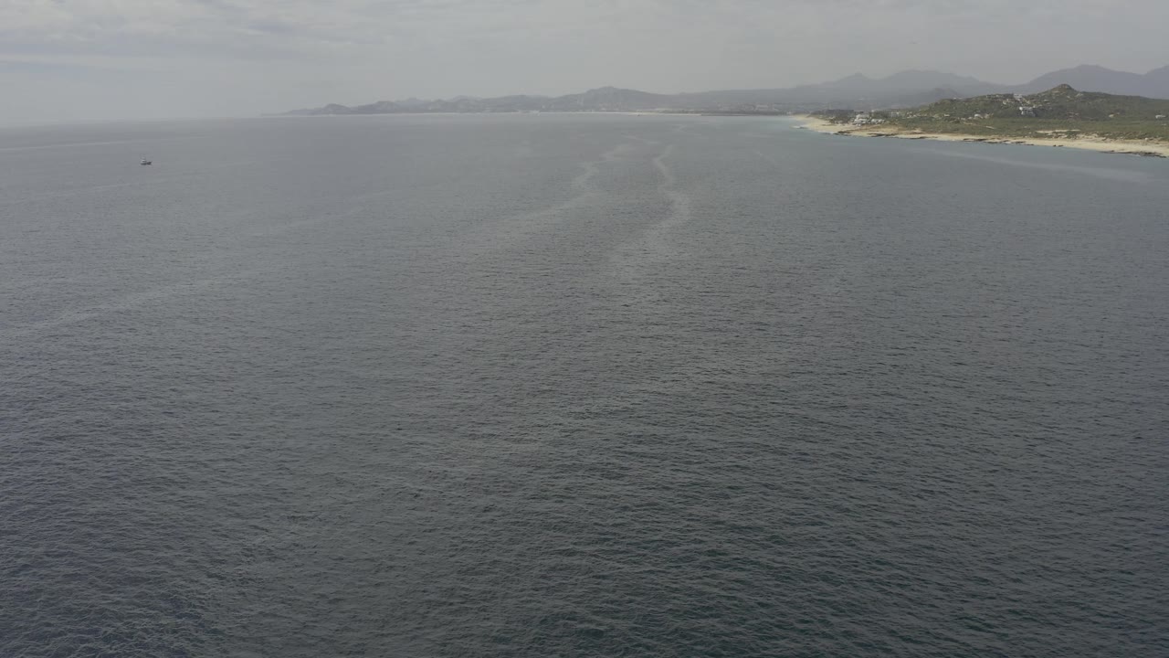 Elevated coastal frame in Baja California Sur showing broad ocean foreground, sandy and rocky shore at upper right, distant hills, hazy daylight, and no visible people.