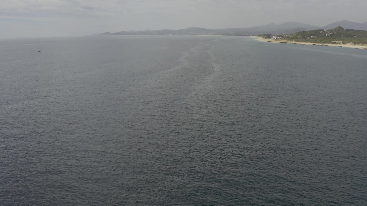 Mid-clip aerial frame over calm Baja California Sur water, with a small dark object left of center, sandy coast to the right, and hazy hills beyond, no visible people.