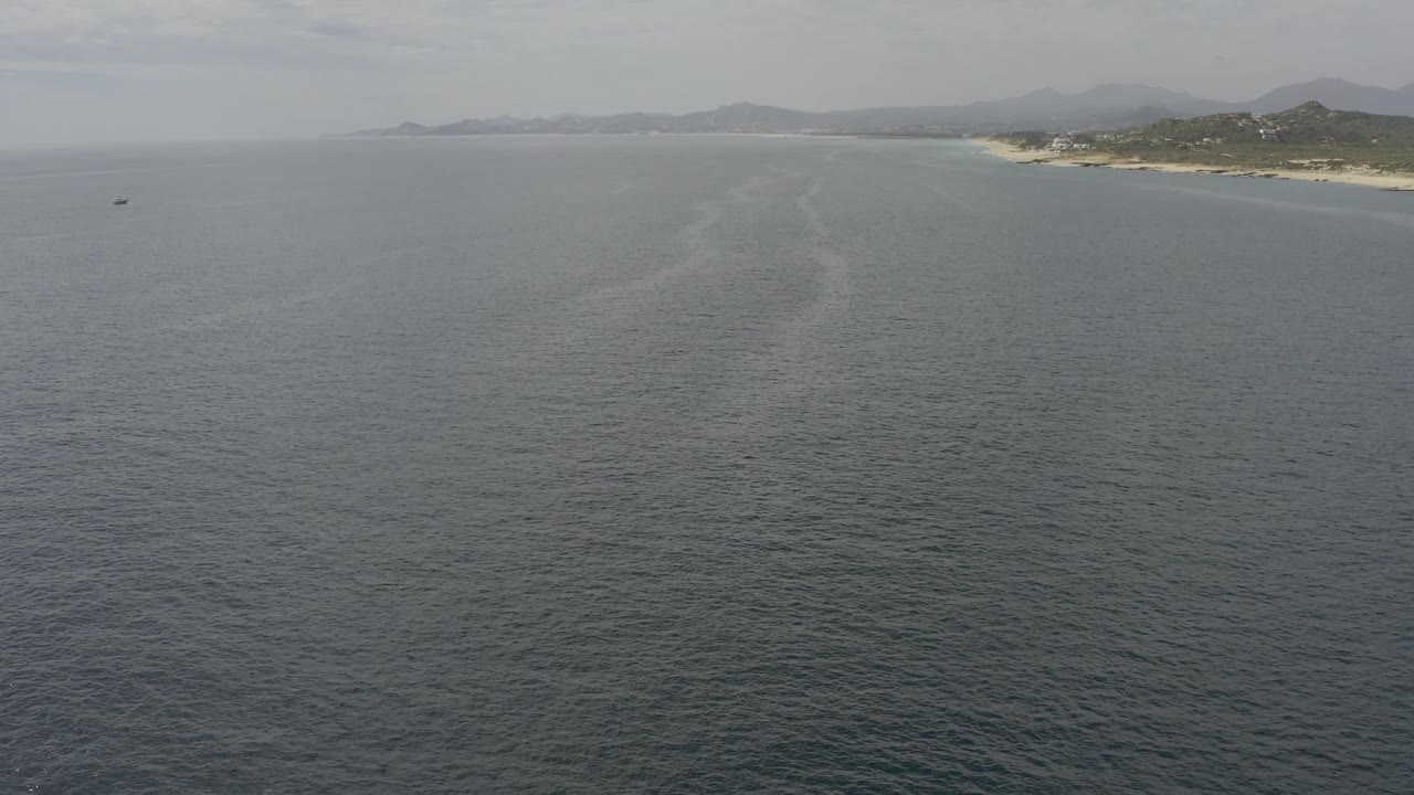 Baja California Sur coastline sits high in frame as gray-blue water fills the foreground and a faint wake line runs toward shore, no visible people.