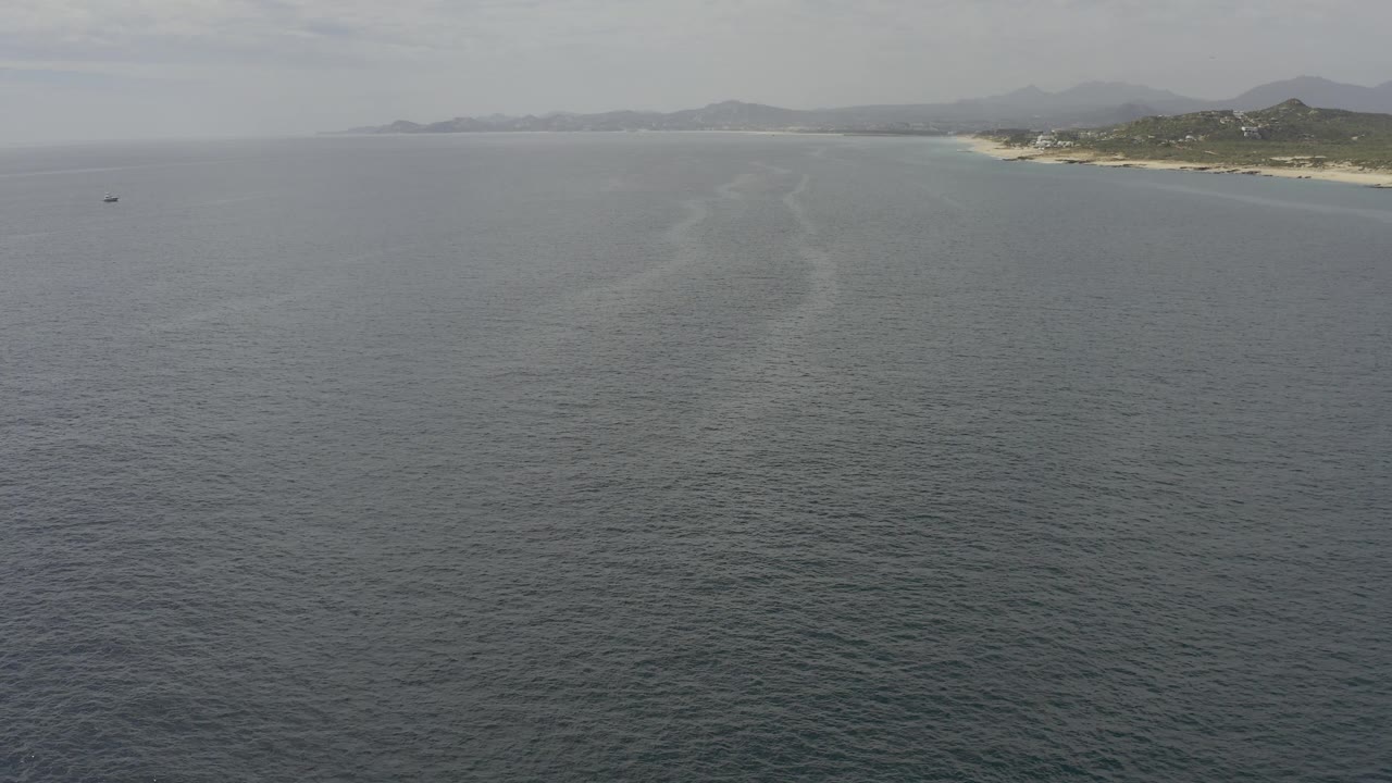 Drone-height view over calm Baja California Sur water with the shoreline angled across the upper right and a small dark boat far left, no visible people.