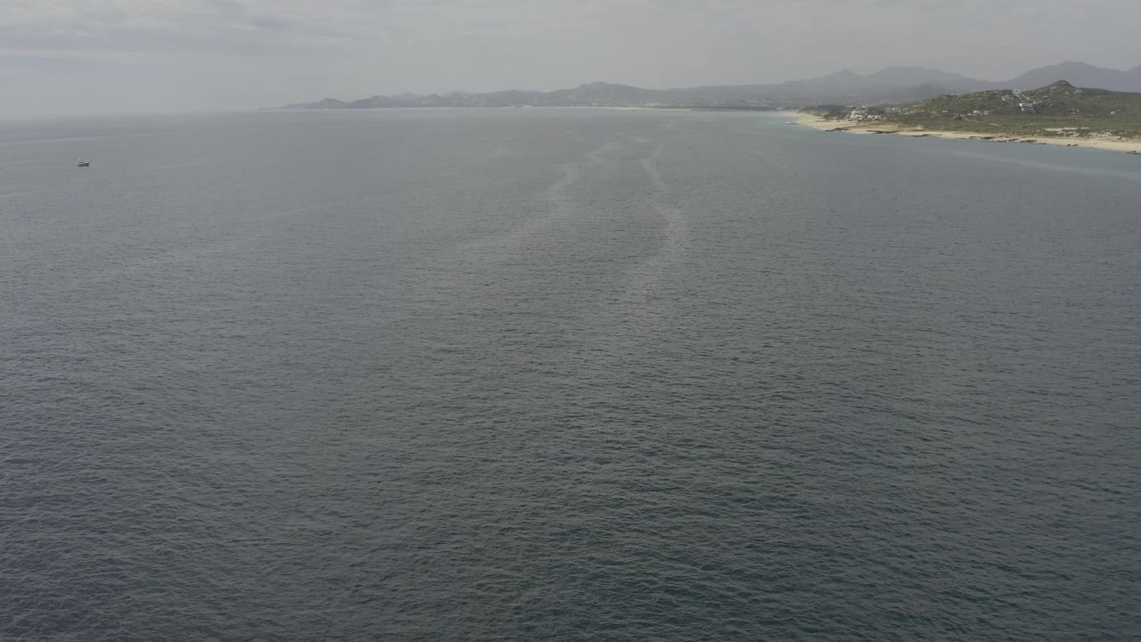 A broad Baja California Sur coastal view holds gray-blue water in the foreground, a faint diagonal water mark, beach at right, and hazy mountains beyond, no visible people.
