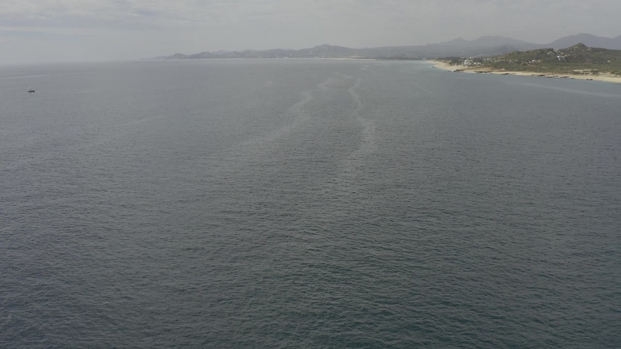 Opening aerial frame over Baja California Sur water, with a broad gray-blue ocean foreground, sandy coastline near the top right, hazy mountains in the distance, and no visible people.