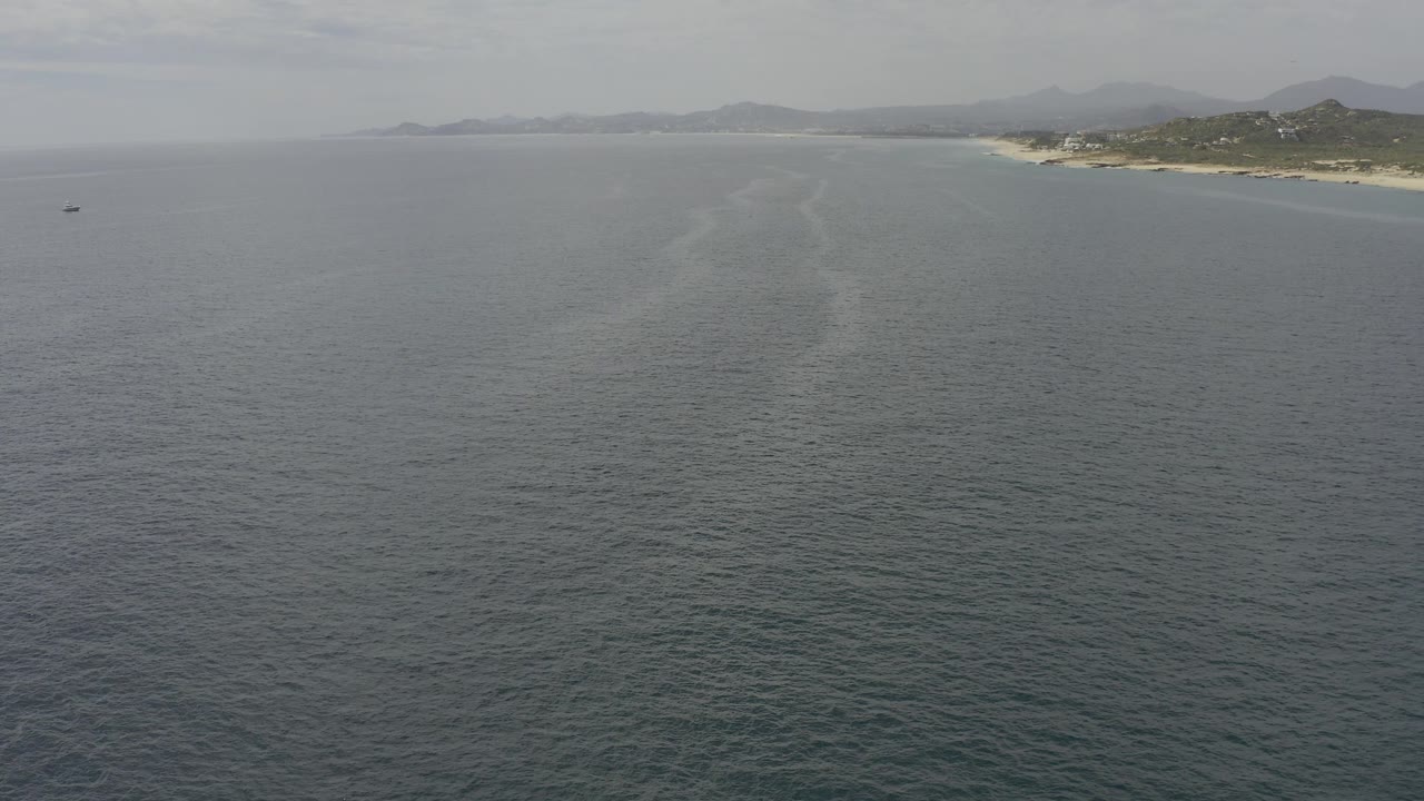 Aerial ocean surface with a pale wake line leading toward a distant beach and hills.