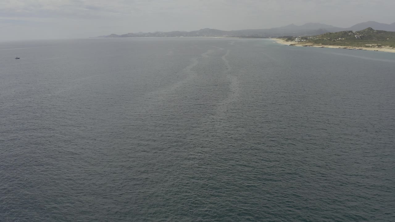 Mid-clip aerial view in Baja California Sur, showing calm gray water, a faint diagonal water trail, sandy shore along the upper right, muted hills, hazy mountains, and no visible people.