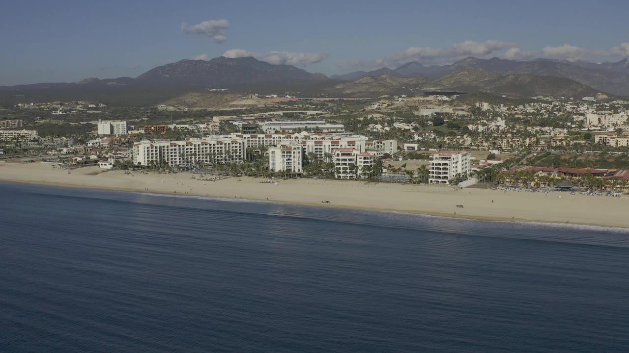 Aerial view in Baja California Sur, Mexico with calm dark water across the lower frame, a narrow surf line, white buildings along the beach, inland streets, mountains, and no visible people.