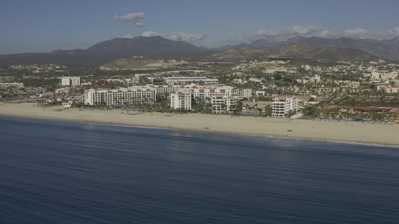 Baja California Sur aerial frame showing a broad ocean foreground, small waves at the shore, clustered white beachfront structures, a low town grid, mountain ridges, scattered clouds, and no visible people.