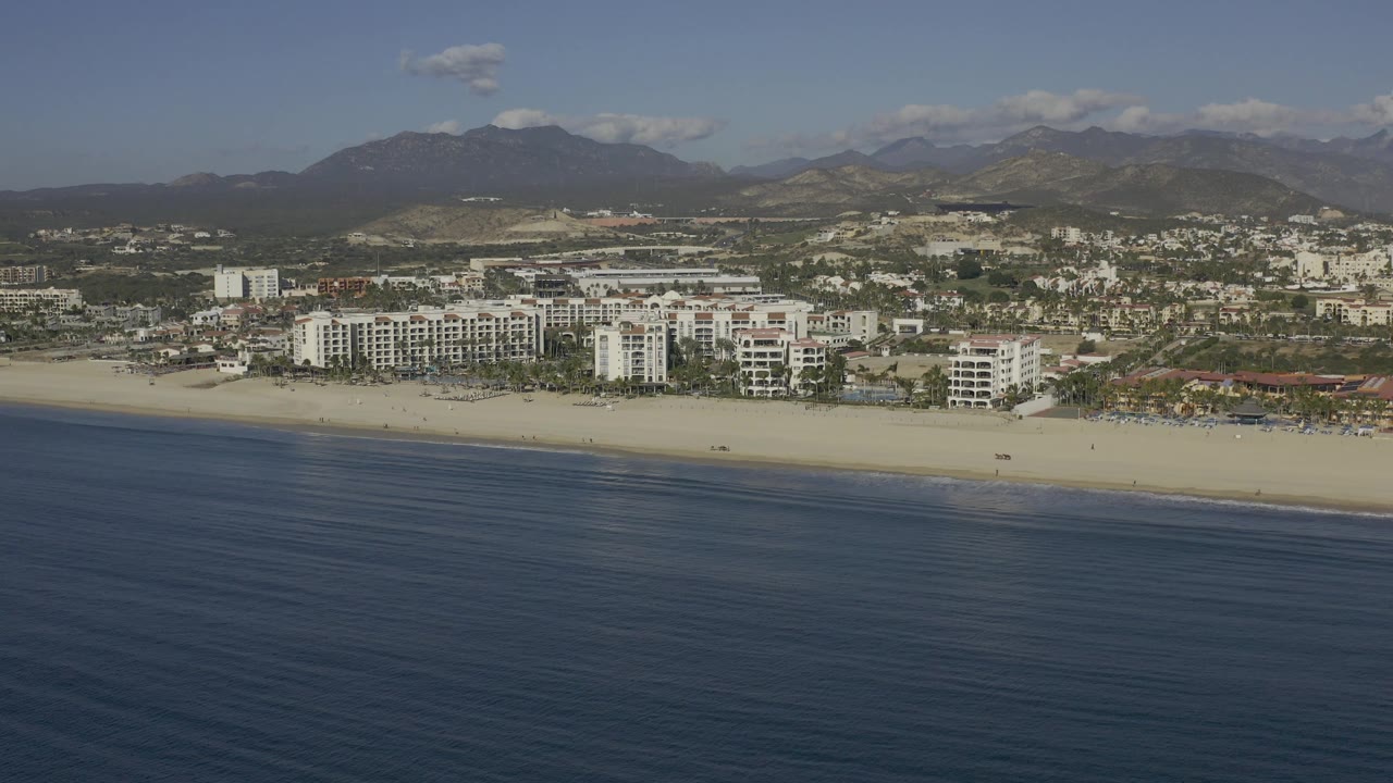 Aerial frame from Baja California Sur, Mexico with darker water in the foreground, a pale beach band, white multi-story buildings near shore, inland development, hazy mountains, and no visible people.