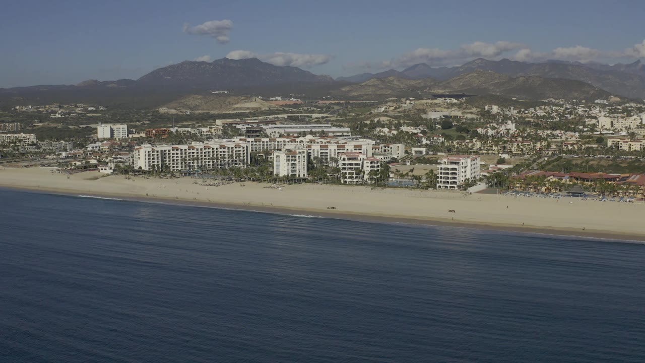 Baja California Sur coastal aerial showing dark blue water below, a thin line of sand, clustered white beachfront buildings, inland roads and low development, mountains, and no visible people.