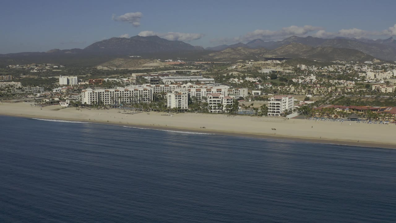 Mid-clip aerial view in Baja California Sur with calm-looking blue ocean across the lower frame, pale sand, low and mid-rise beachfront buildings, inland hills, mountains, and no visible people.