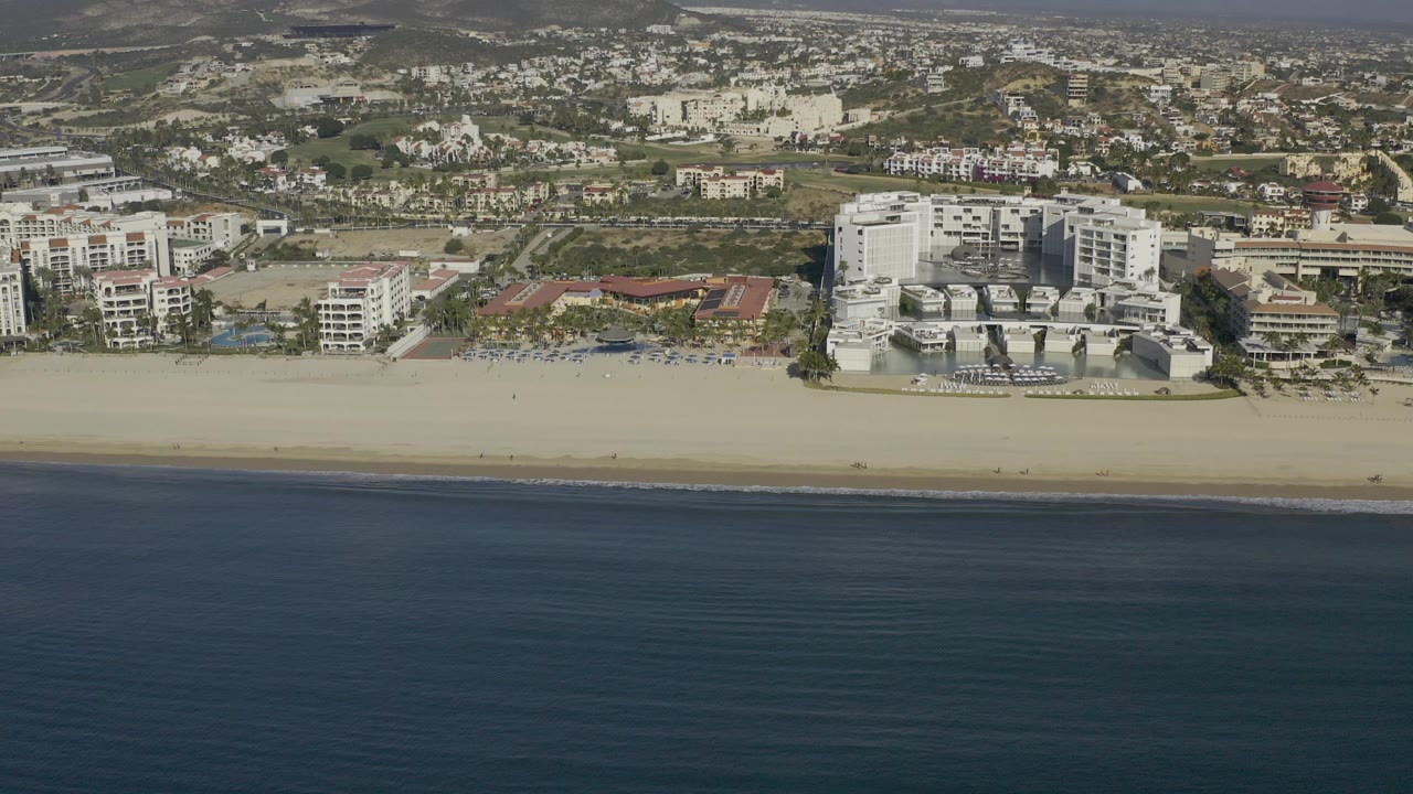 Aerial frame over Baja California Sur, Mexico, showing dark blue ocean, a pale sand beach, beachfront hotel buildings, inland roads, and low hills beyond.