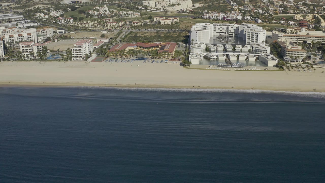 The second frame keeps the Baja California Sur shoreline centered, with calm dark water, an empty-looking sand strip, a large white resort-style complex, and no visible people.