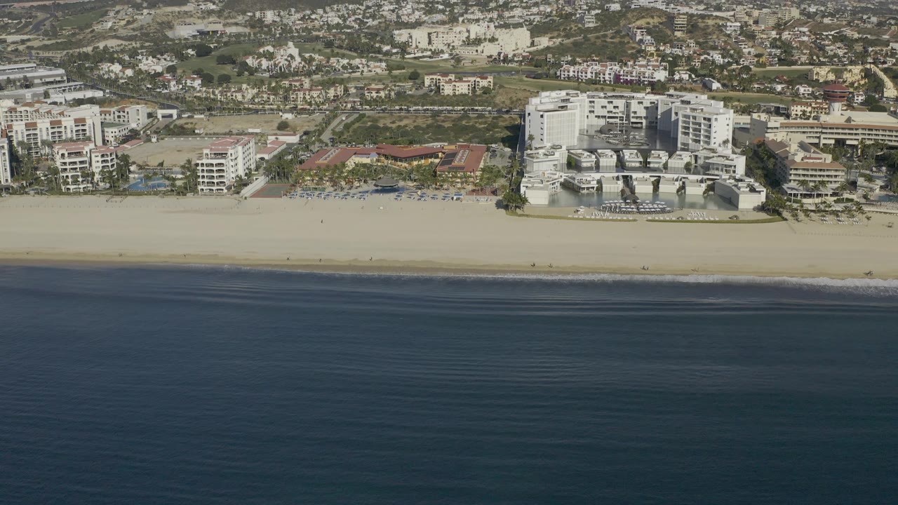This Baja California Sur frame shows the coastline from above, with a dark ocean band below, pale sand, clustered beachfront buildings, dry inland hills, and no visible people.