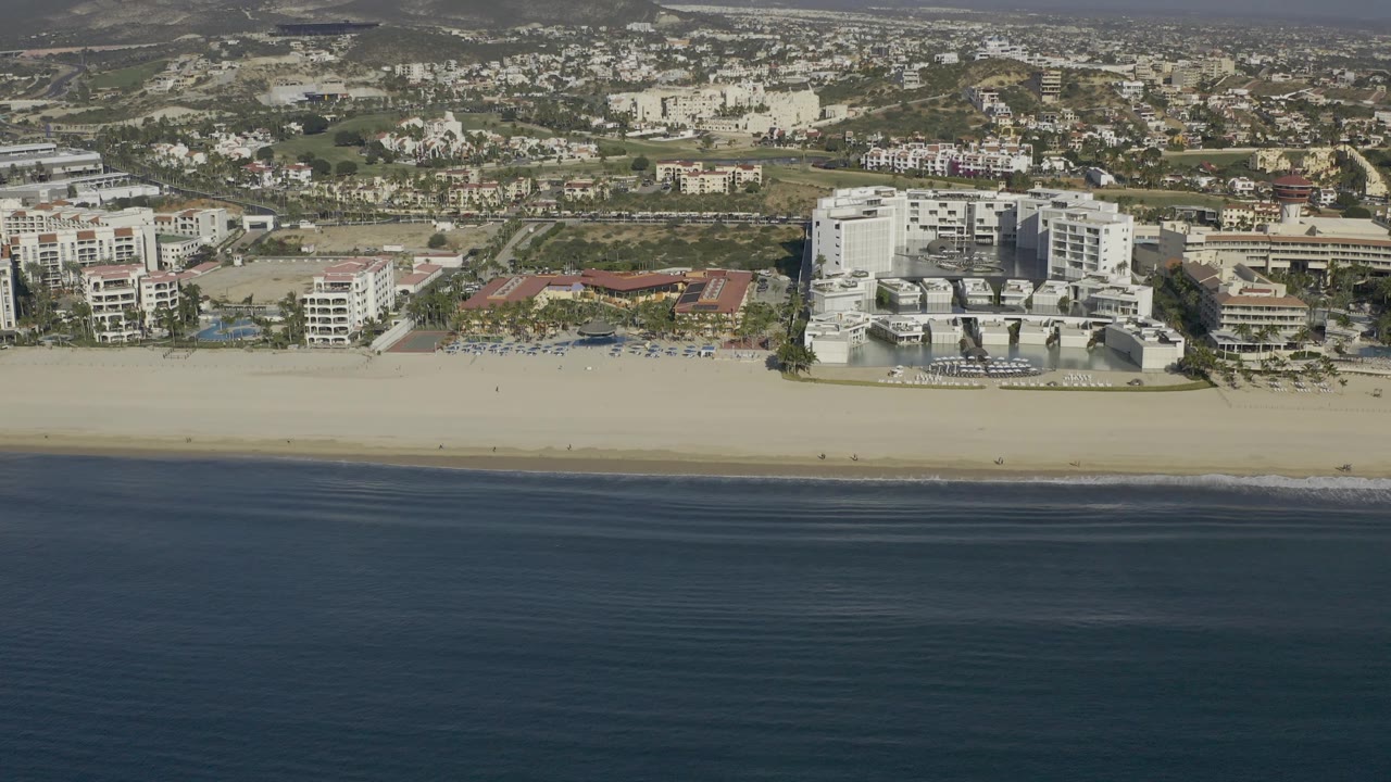 At 0:03, the aerial view shows calm blue water, a long empty-looking beach, a white beachfront complex, surrounding resort or residential buildings, and no visible people in Baja California Sur.