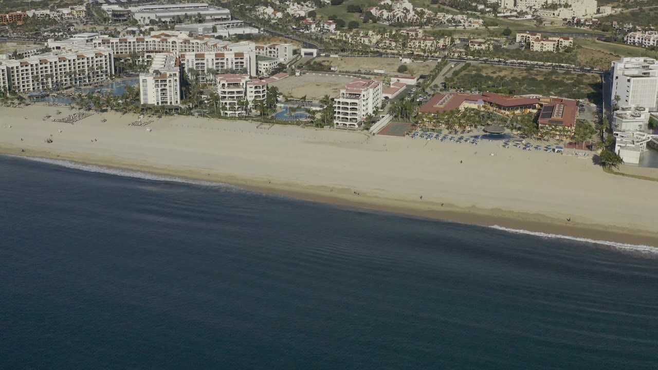 Aerial frame over Baja California Sur, Mexico showing dark blue ocean below, a tan beach through the center, white beachfront buildings beyond the sand, and no visible people.