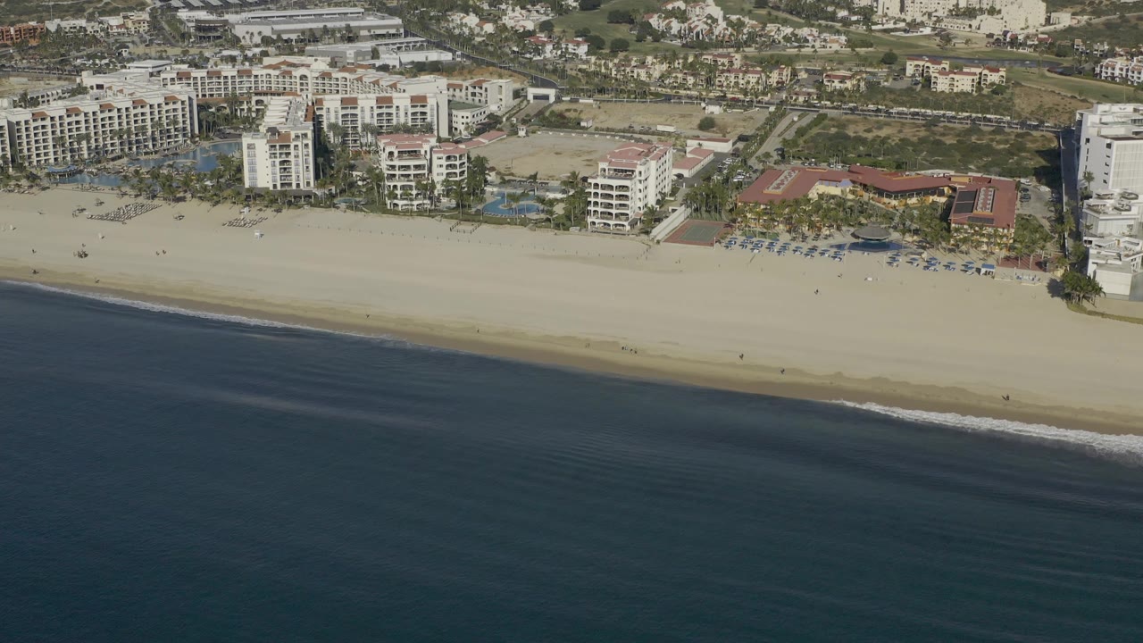 Oblique aerial frame over Baja California Sur, Mexico showing the beach running horizontally, ocean in the lower half, clustered white buildings near the sand, and no visible people.