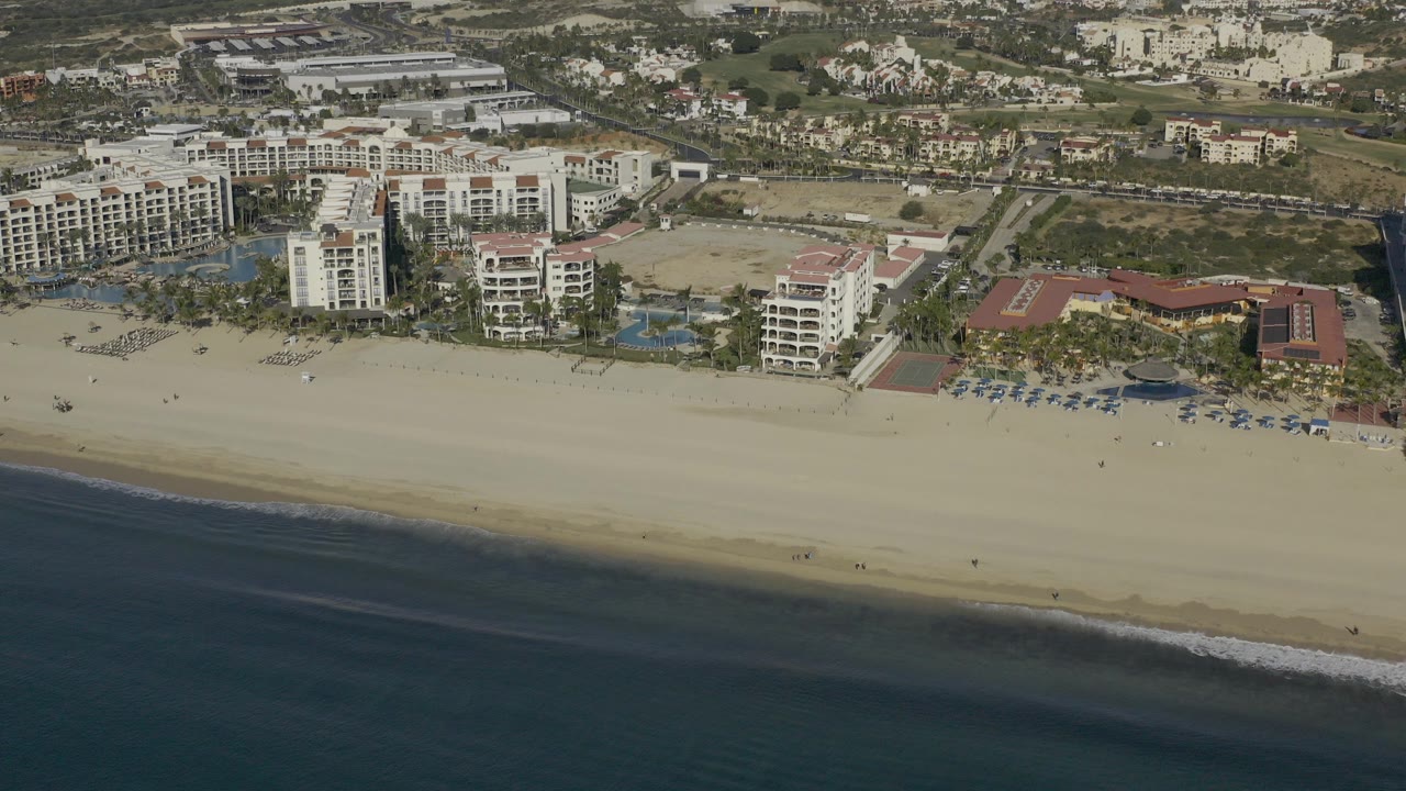 Aerial daytime view in Baja California Sur Mexico showing dark ocean water, wide sand, beachfront hotel buildings, pools, and inland roads with no visible people.