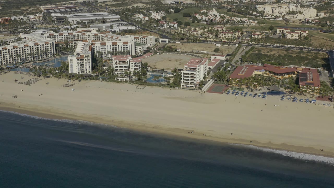 Aerial frame in Baja California Sur Mexico with the ocean along the lower edge, a broad pale beach, clustered hotel buildings, blue pools, and roads behind them with no visible people.