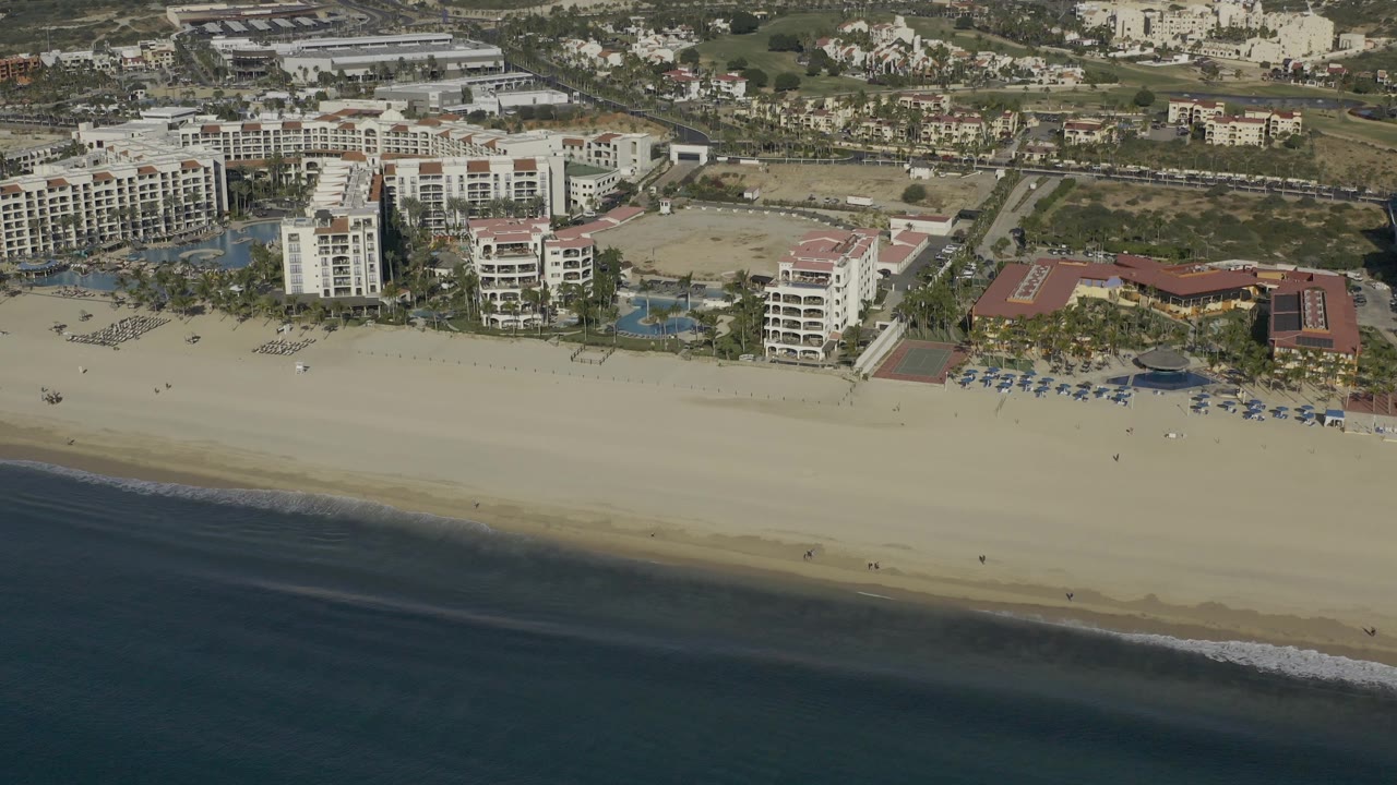 Daytime aerial view in Baja California Sur Mexico showing the resort buildings slightly shifted across frame, dark water in front, a wide beach, pools, and inland lots with no visible people.