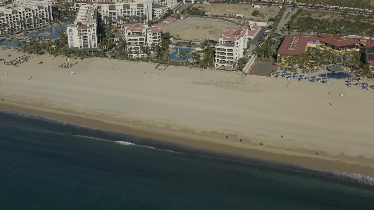 High aerial view in Baja California Sur, Mexico, with hotel buildings, pools, palms, pale sand, and dark ocean water below, with no visible people identifiable.