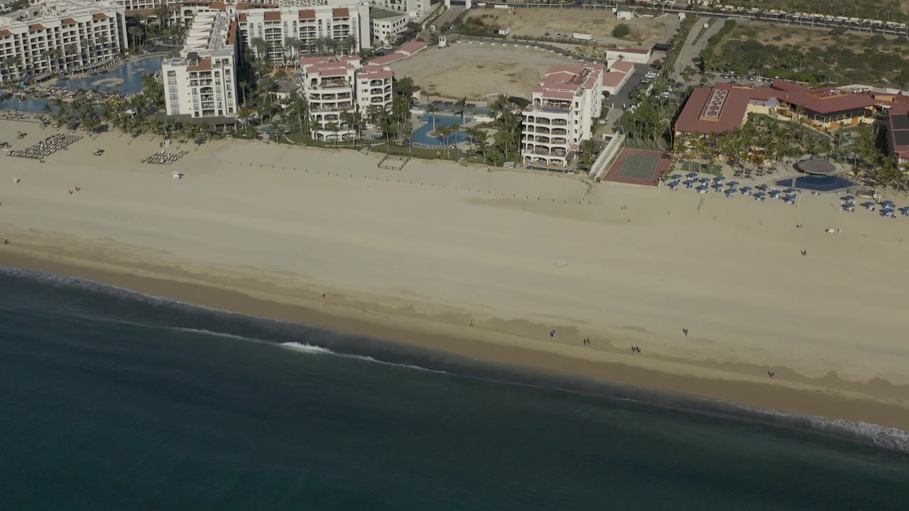 Aerial frame over Baja California Sur shoreline showing resort pools and buildings above a broad empty-looking beach, with a thin wave line meeting the sand and no visible people identifiable.