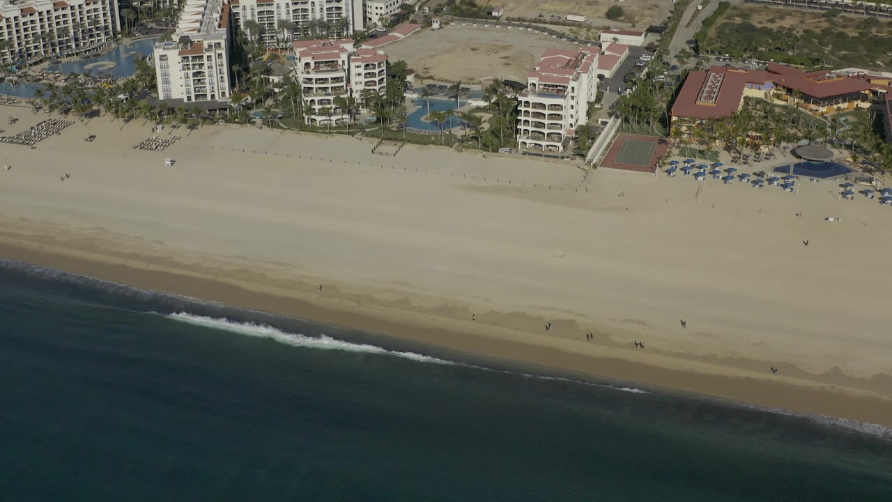 High oblique view of Baja California Sur beach where hotel grounds and rows of umbrellas sit above a wide sand strip, with a brighter surf line entering from the lower right and no visible people identifiable.