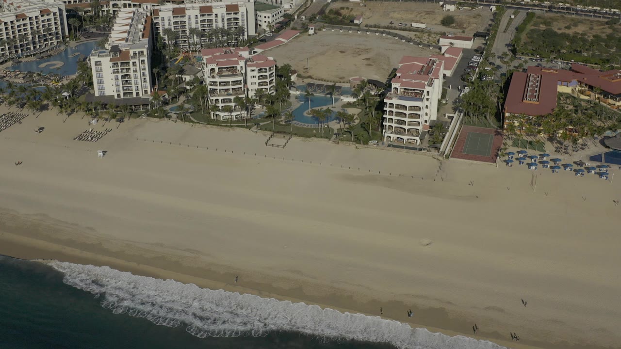 High aerial view in Baja California Sur showing beachfront hotel buildings, pools, palms, broad sand, and a strip of surf at the bottom, with no visible people.