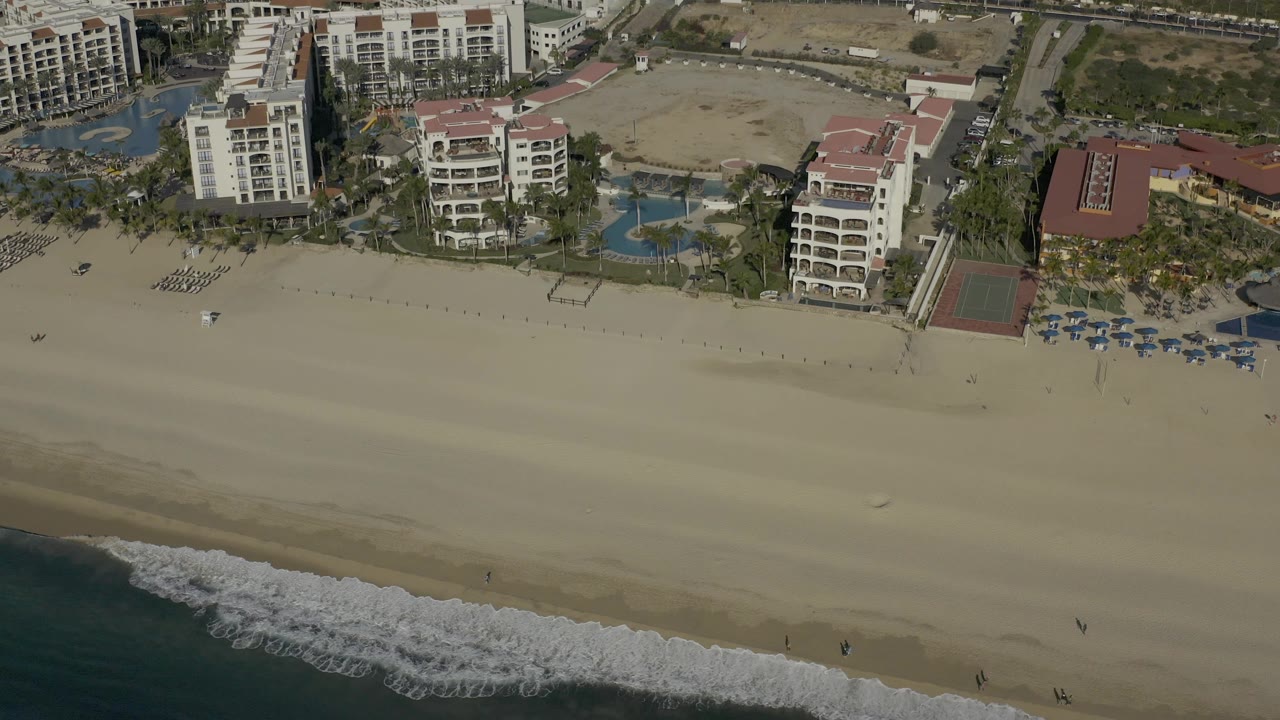 Aerial frame in Baja California Sur with the surf line shifted across the lower left, open sand in the center, and hotel buildings, pools, palms, and roads above, with no visible people.