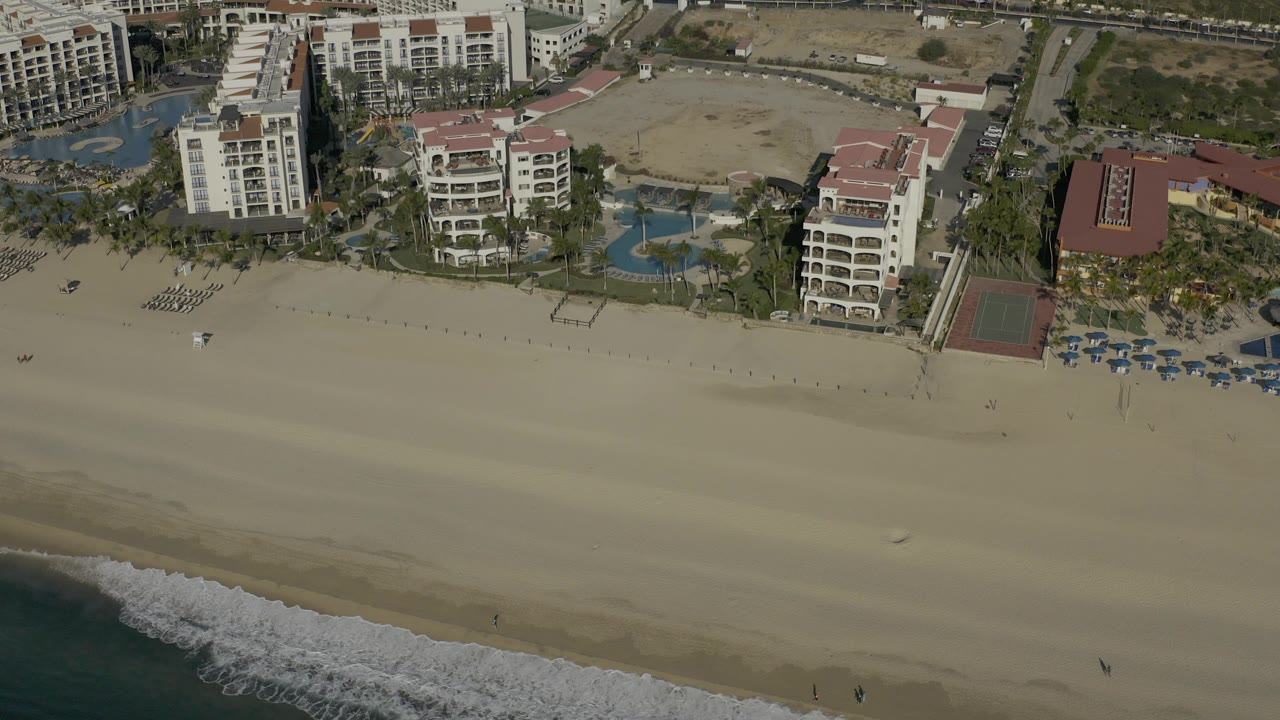 High oblique aerial view in Baja California Sur with foamy surf entering from the lower left, a wide beach across the middle, and clustered hotel buildings and pools at the top, with no visible people.