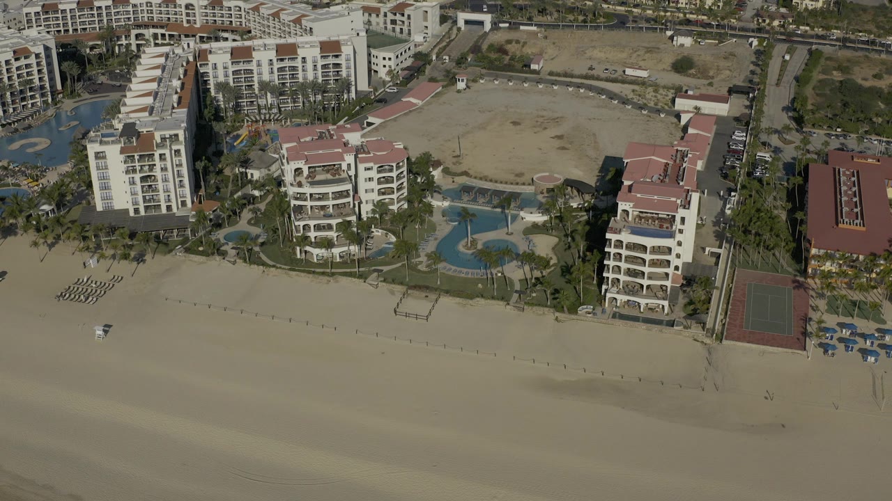 Opening aerial frame over a Baja California Sur beachfront resort, with cream buildings, blue pools, a tennis court, sandy beach, and no visible people.