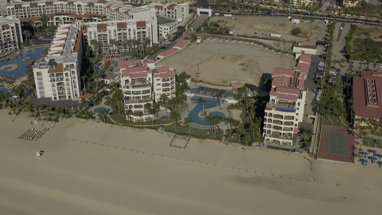 Baja California Sur aerial view with the resort centered, blue pools and palms below the buildings, beach loungers near the sand, a tennis court at right, and no visible people.