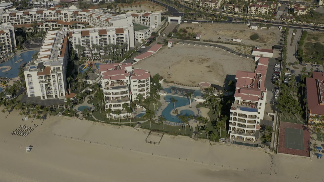 Aerial resort view with beach, pools, multiple buildings, and inland road.