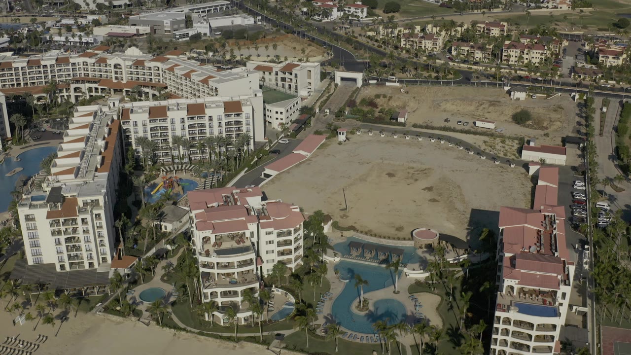 Drone view of white buildings, curved pool areas, palms, and nearby roads.