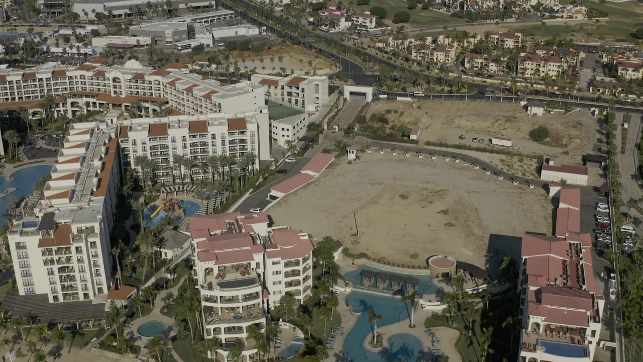 High aerial frame over Baja California Sur, Mexico showing red-roofed resort buildings, rows of palms, blue pools, a large vacant sandy lot, roads, and no visible people.