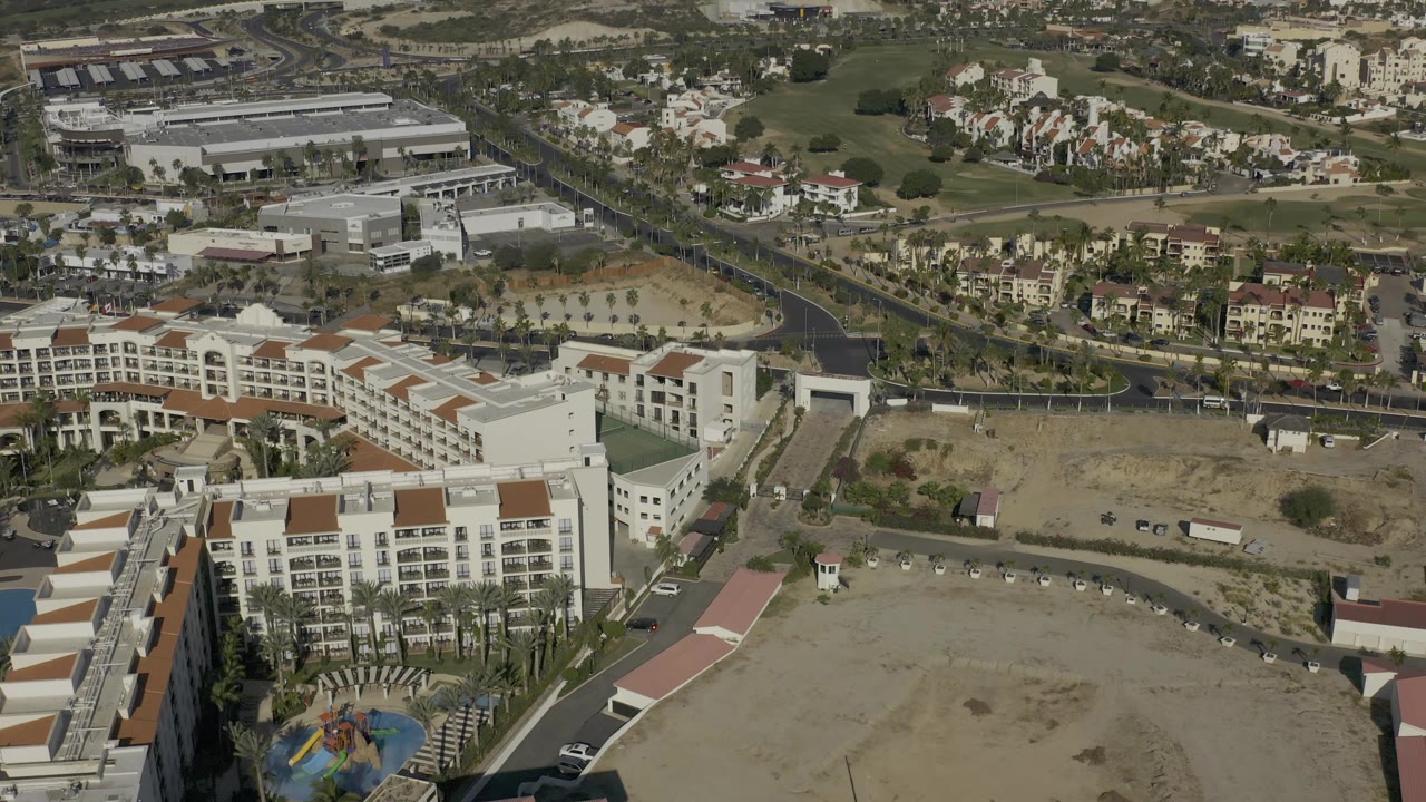 Aerial frame in Baja California Sur, Mexico, with a large white hotel block in the foreground, a dry lot and roadway in the center, and residential clusters near green fairways beyond, with no visible people.
