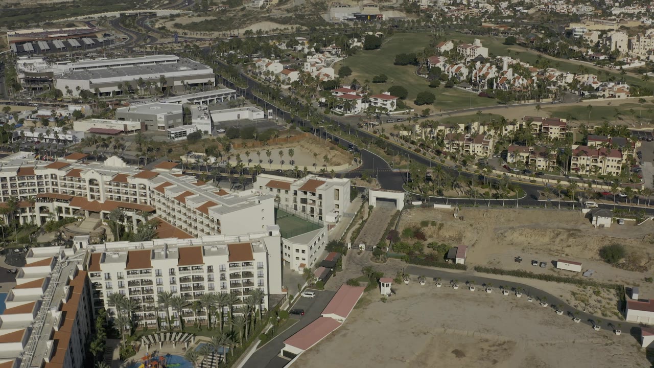 High oblique view in Baja California Sur, Mexico, looking across resort roofs and a curved access road toward golf-course grass, low-rise buildings, and dry hills, with no visible people.