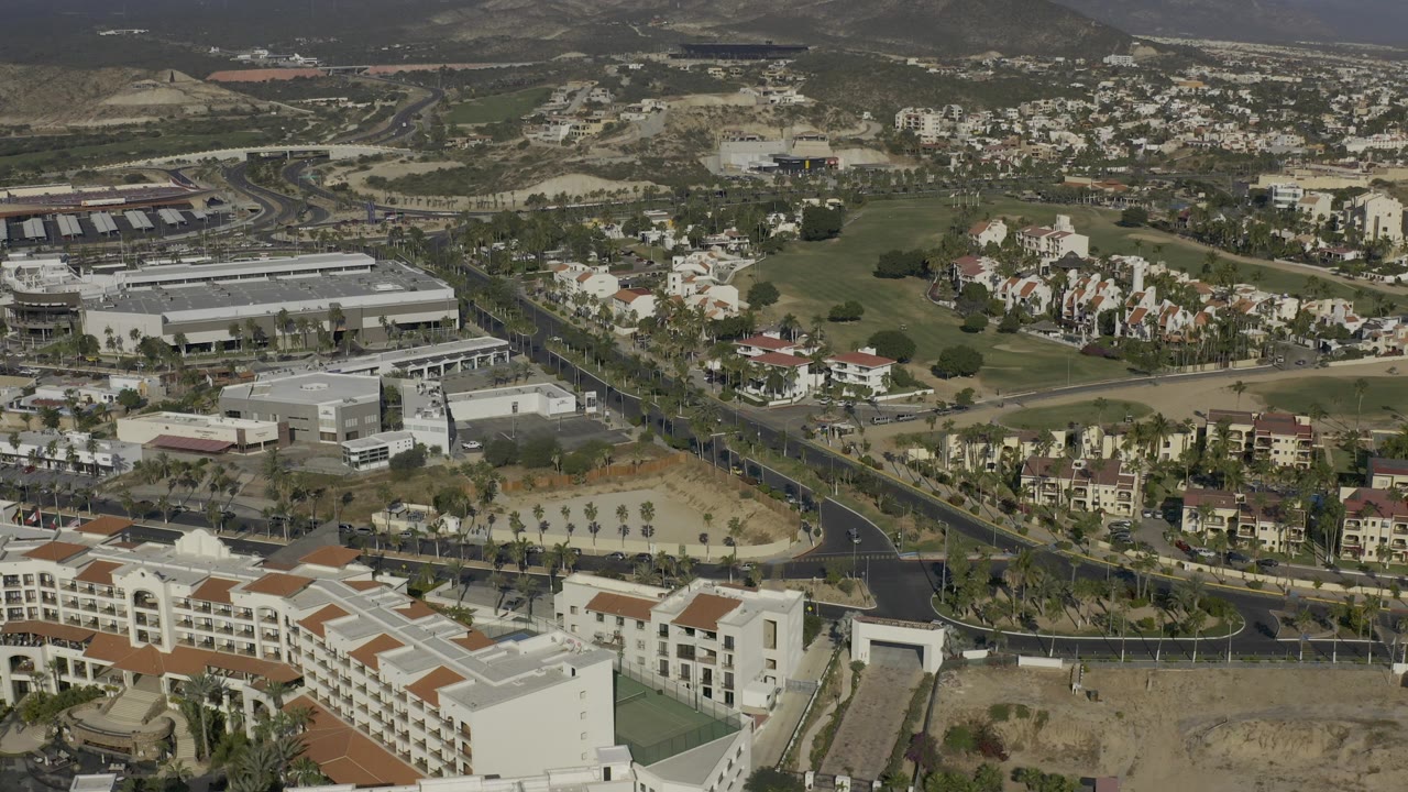 Opening aerial frame over a Baja California Sur town with white buildings in the foreground, curving roads, clustered homes, green landscaped areas, distant mountains, and no visible people.