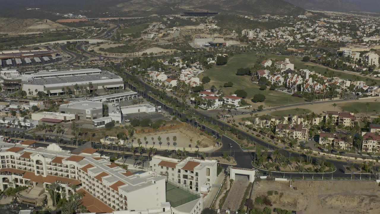 Early aerial frame in Baja California Sur showing a large light-roofed building near the center, surrounding homes, palm-lined roads, dry terrain, distant hills, and no visible people.